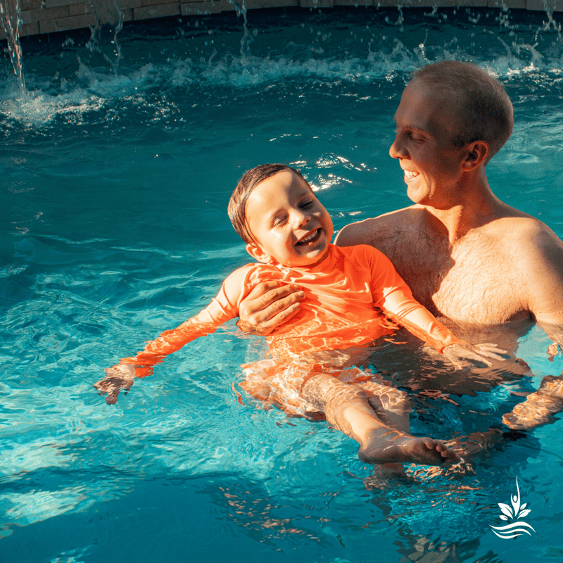 A joyful father and son play in their pool, with the father holding his preschool-aged son in his arms. The son has a big smile on his face and a relaxed pose, arms wide open, as soft waterfalls cascade in the background, creating a scene of familial bliss and outdoor fun.
