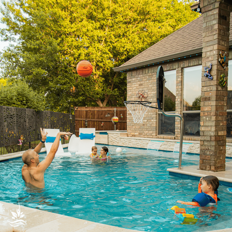 A family of four enjoys a playful moment by the pool. The mother, holding her daughter, watches as the father, with a determined leap, makes a successful jump shot at the basketball hoop, much to the delight of their son and the gentle applause of waterfalls in the background.