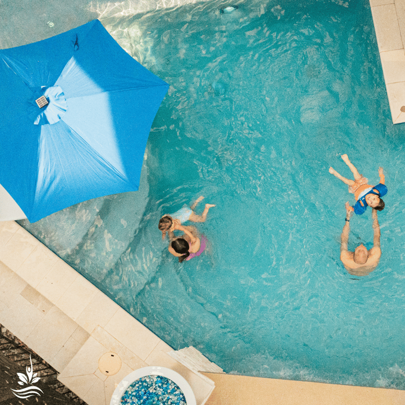 An aerial view captures our family of four in their custom swimming pool, immersed in playful moments. Their young son, aware of the camera above, looks up with a beaming smile. The big blue pool umbrella, unfolded and prominent, adds to the cheerful scene.