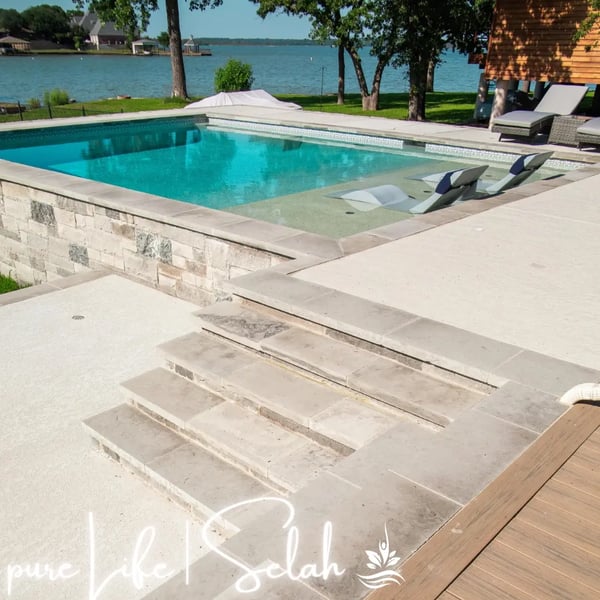Oblique view of a custom-designed, elevated pool area by Selah Pools in Argyle, Texas, featuring a pool, tanning ledge, semi-submerged lounge chairs, integrated concrete steps, and travertine walkways. A luxurious waterfront view is visible in the background. The scene is crafted in harmony with the land.