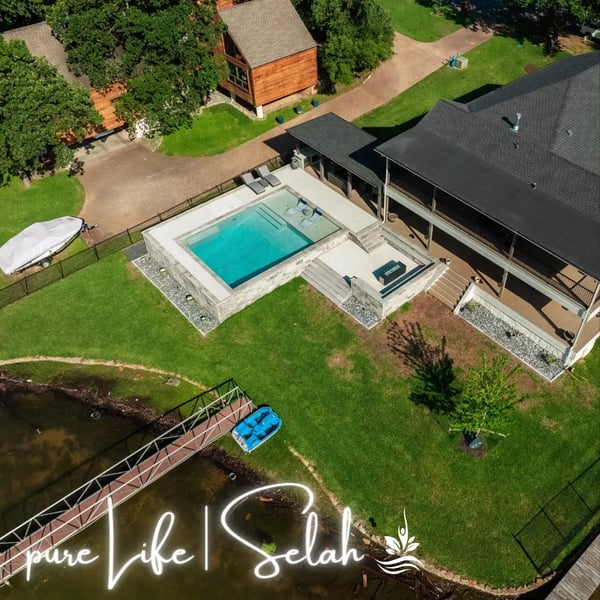 Aerial view of a complete outdoor scene by Selah Pools, featuring a custom swimming pool, a sunken conversation pit with a linear edge firepit, a custom patio extension, and a shade structure, all elevated above the waterfront.
