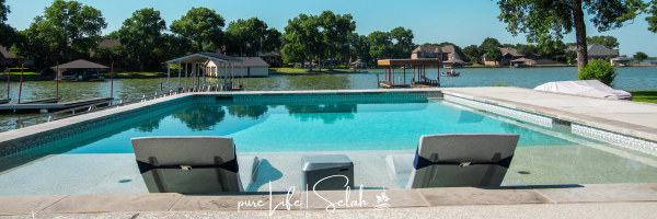 An inviting backyard oasis featuring a clear blue swimming pool with two lounge chairs, surrounded by a well-maintained lawn, overlooking a peaceful lake with docks and boats, under a clear sky. This serene setting encapsulates the tranquility and beauty of a custom outdoor environment designed by Selah Pools.